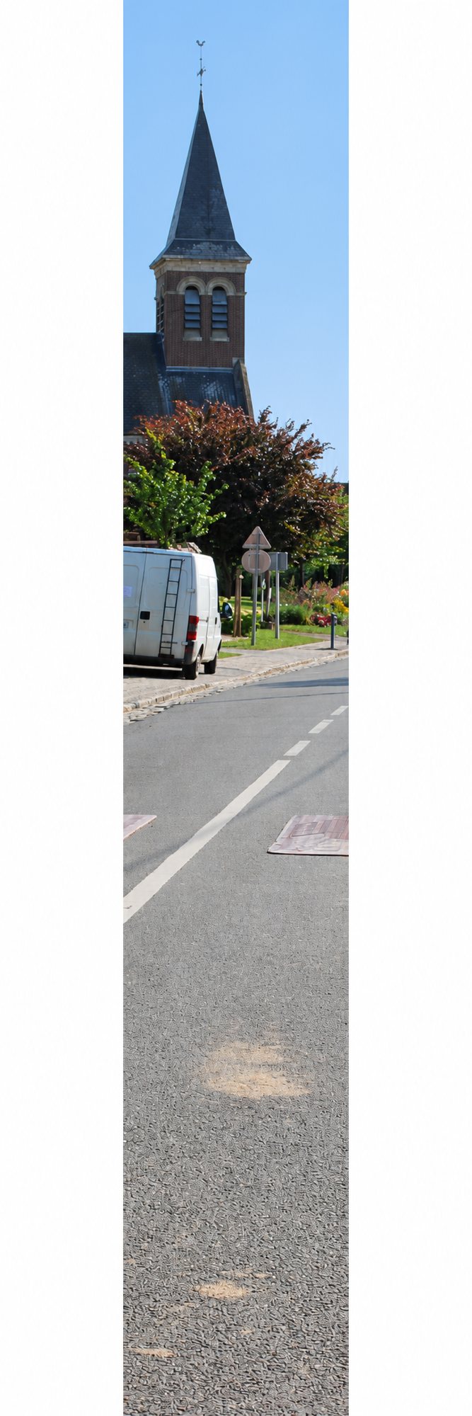 Route avec ralentisseur et camionnette blanche, près d’un village dominé par un clocher en brique